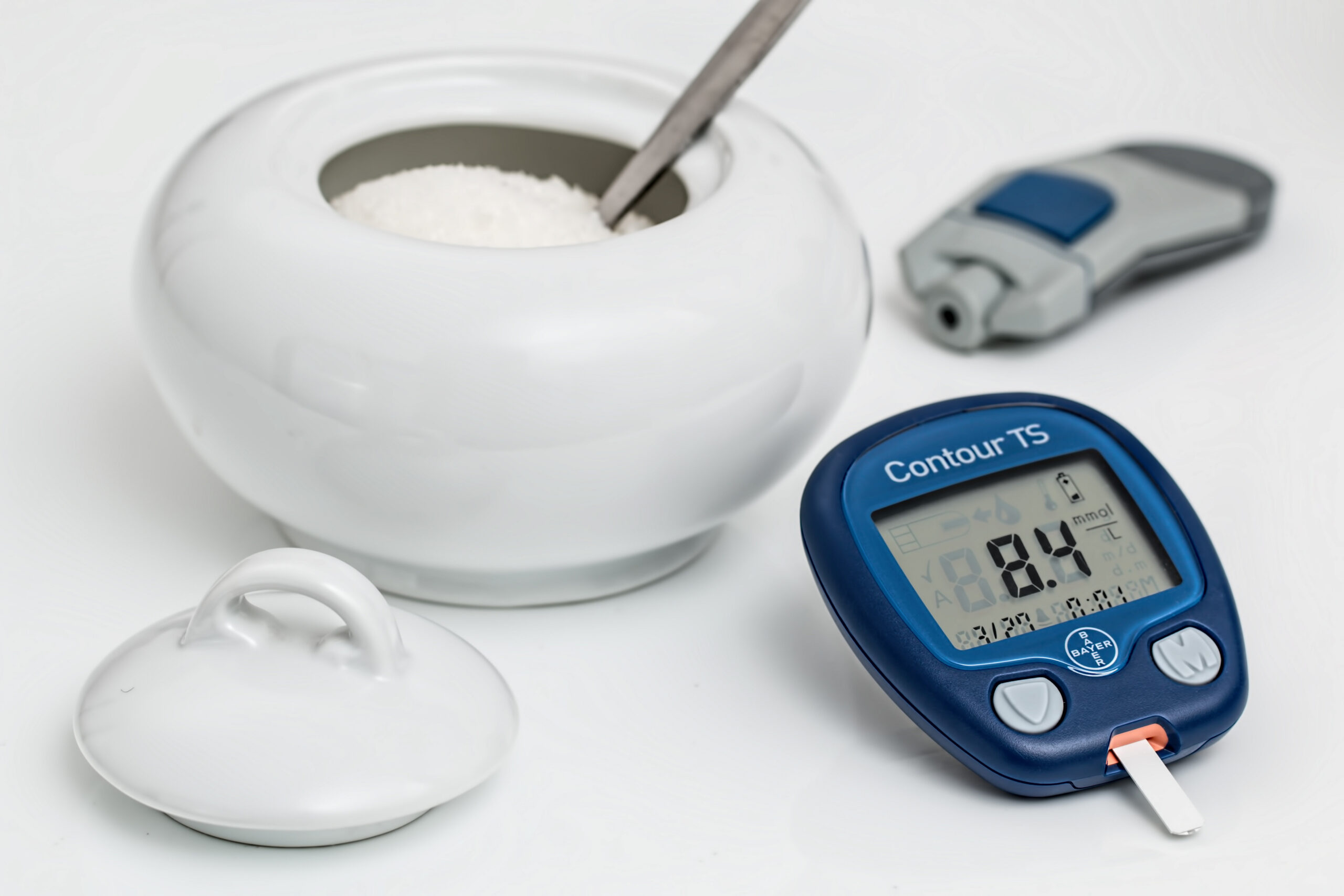 Glukomer s nameranou hodnotou 8,4 mmol/l vedľa misky s cukrom a ďalšieho glukomera.
